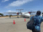 Man with backpack approaches white airplane with red engines on a runway. Traffic cones and a security person are visible. Clear blue sky. Arrival at Manada airport, Kenya opposite Lamu Island.