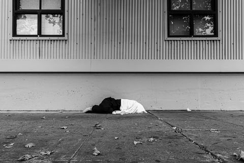 Eine Obdachlose Person liegt auf der Strasse, der Oberkörper der Person ist mit einer Decke zugedeckt, im Hintergrund eine einfache Hausfassade mit zwei Fenstern 