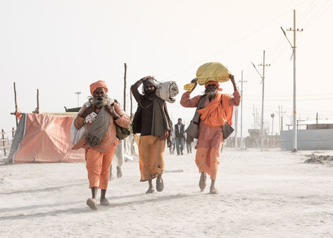 Drei Sadhus in orangen Gewändern tragen Bündel auf Schultern und Köpfen durch staubige Wege
