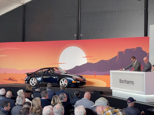 Bidders seated in front of a Bonhams auction stage as a dark blue Porsche 993 crosses the block under a desert sunset backdrop.