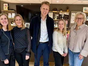 Group of five smiling professionals, three women and two men, standing in a modern office with wooden flooring, cozy furniture, and shelves filled with awards, trophies, framed certificates, and artwork. The individuals are dressed in business casual attire, suggesting a workplace or corporate environment. A large flat-screen TV is mounted on the back wall, contributing to the contemporary office setting.