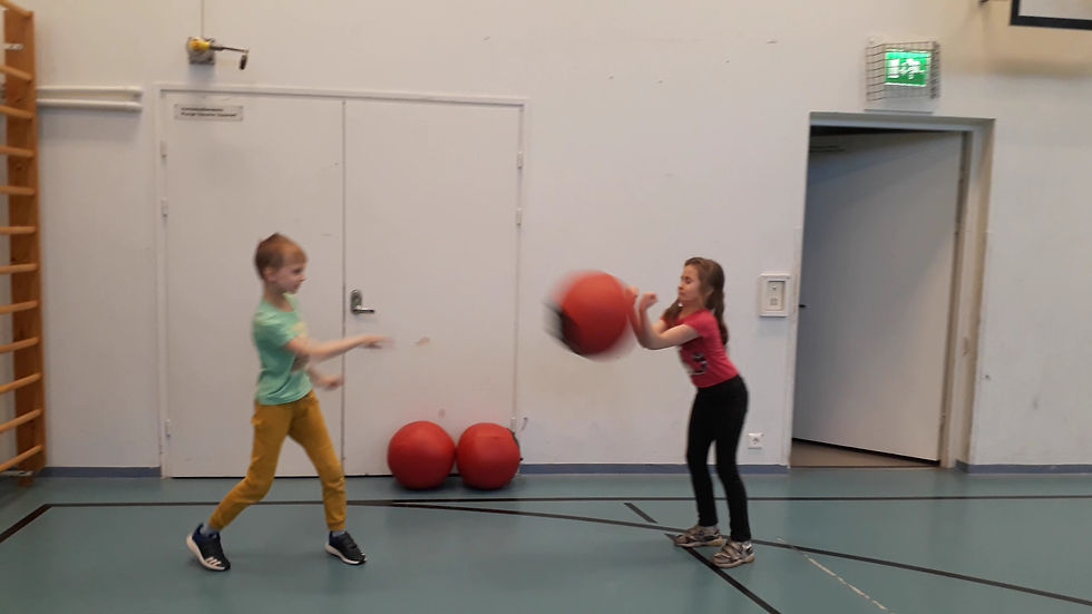 Kaksi lasta heittelee punaista palloa salissa. Poika vihreässä paidassa, tyttö pinkissä. Taustalla valkoiset seinät ja ovi.