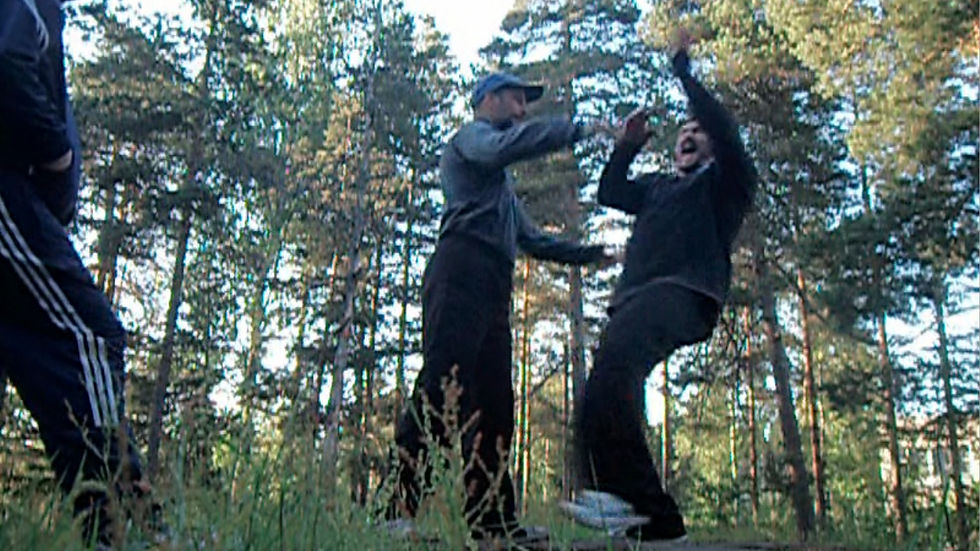 Two men in dark sportswear spar or high-five in a pine forest, with a third person partly visible at left.