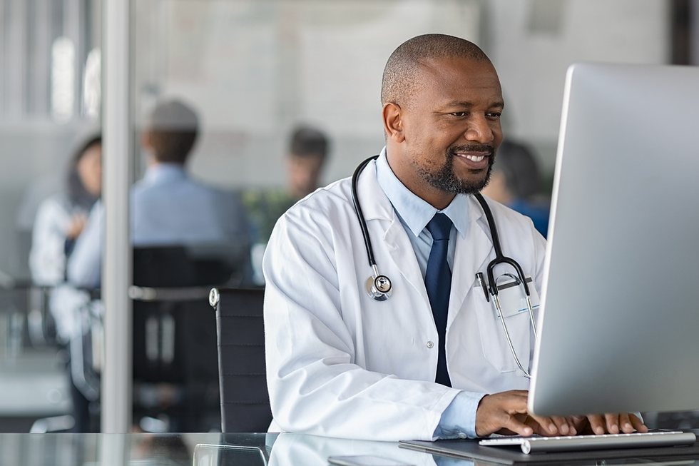 Smiling doctor in a white coat with a stethoscope works on a computer in a modern office. Blurred colleagues in background.