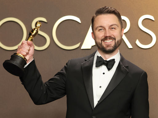 Man in a tuxedo holds a golden statue with the word Oscars behind him.