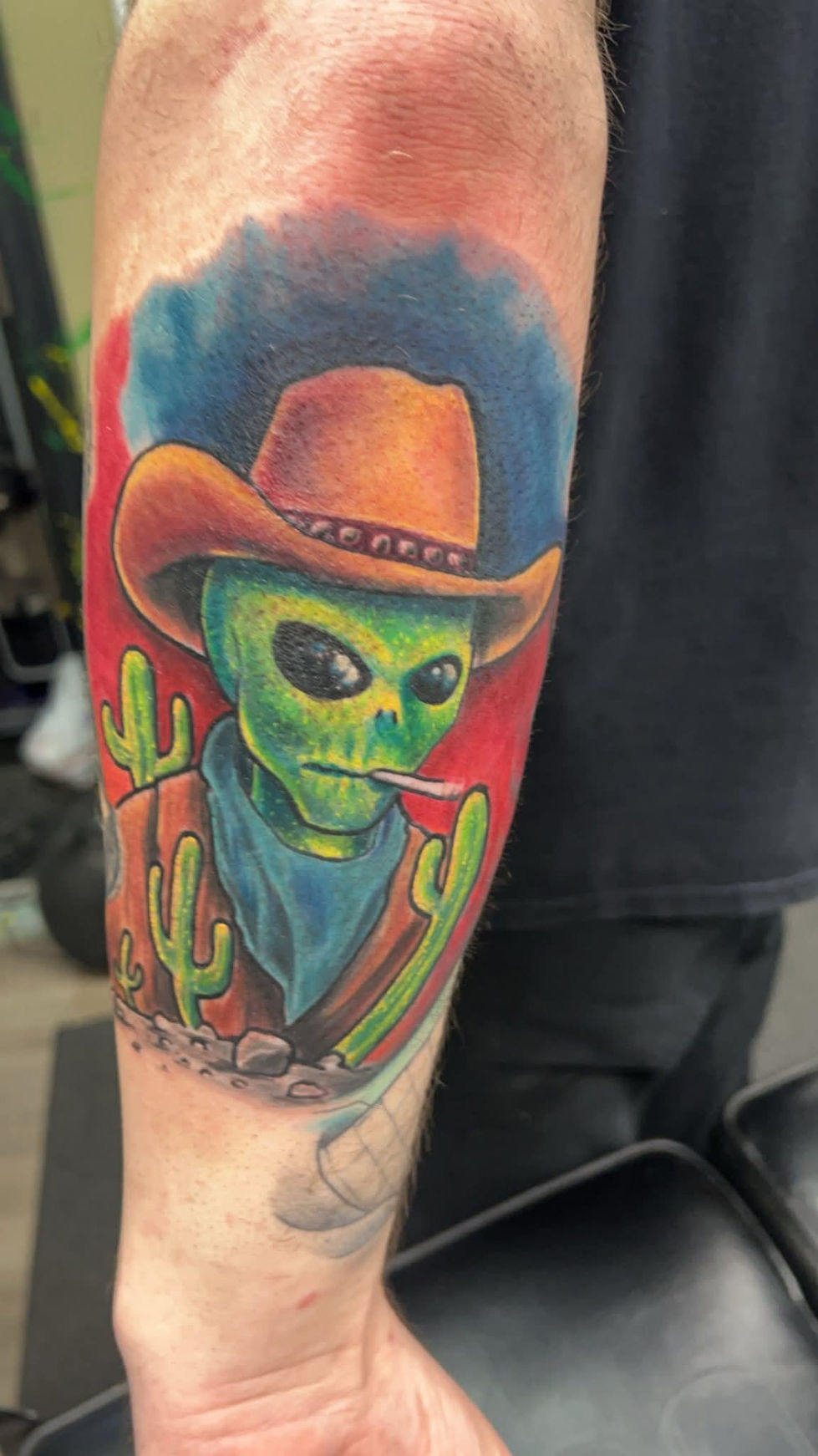 Cowboy alien tattoo, wearing a hat and smoking, with cactus in the background.