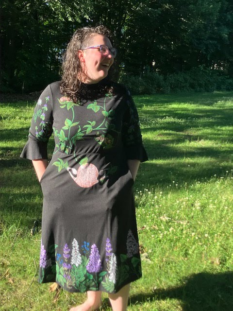 Laughing woman in a nature and anatomy-themed dress stands on sunlit grass, hands in pockets. She's wearing glasses, with trees in the background.