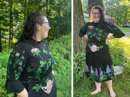 Woman smiling in a black dress with hand-painted floral and anatomy designs stands barefoot on grass by tall trees, with a sunny garden in the background.