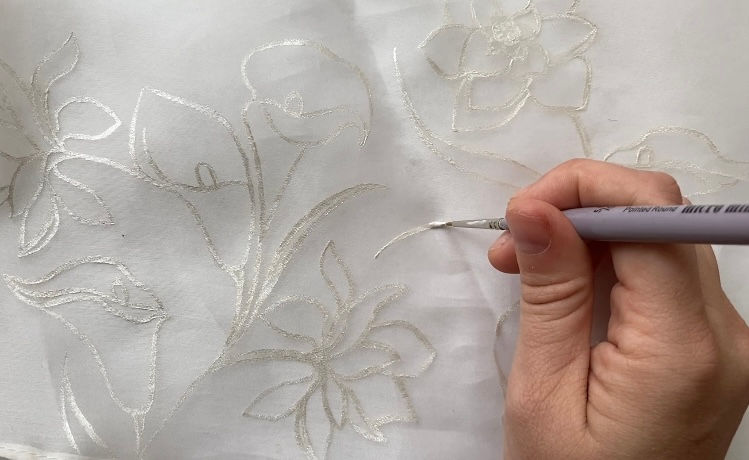 Hand holding a paintbrush creates silvery floral patterns on translucent white fabric, creating elegant designs.