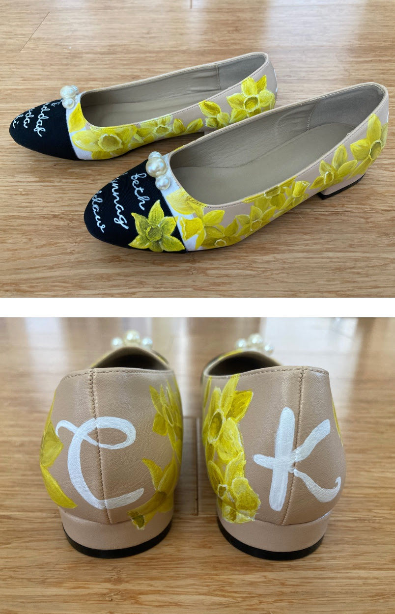 Beige flats with hand-painted yellow flowers and pearl accents on wooden floor. Black toe with white cursive text. Initials "C" and "K" on back.