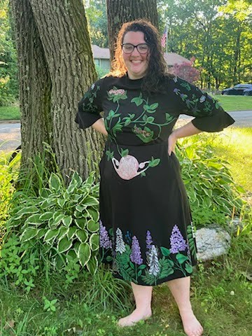 Woman smiling in a black dress with hand-painted floral and anatomy designs stands barefoot on grass by tall trees, with a sunny garden in the background.
