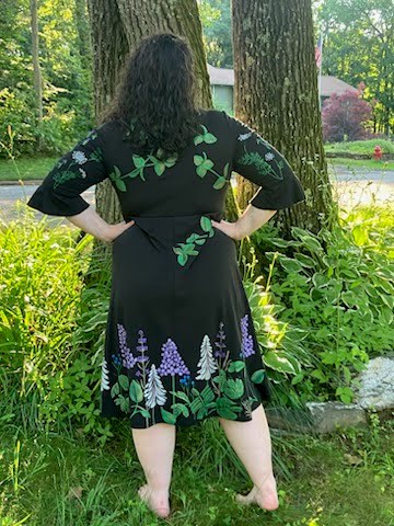 Woman in a black hand-painted floral dress stands barefoot, facing two trees in a lush garden. Bright greenery and a house are visible in the background.