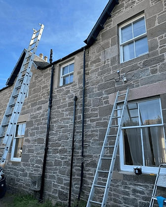 The image shows a two-story stone house with a slate roof, white sash windows, black downpipes, and extension ladders set up against the exterior walls for maintenance work.