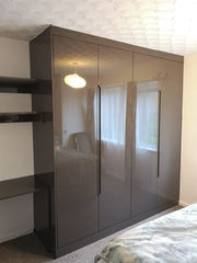 A modern bedroom features a glossy brown wardrobe with sleek handles, a built-in desk, and shelves