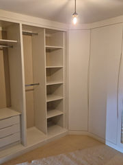 A beige, empty corner wardrobe with open shelves, hanging rods, and a closed cabinet door