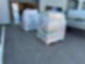 Two large pallets wrapped in transparent plastic sit outside an open warehouse with visible stock and a delivery truck