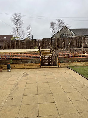 A scene with a paved patio. Wooden steps lead up through terraced earth to a fence