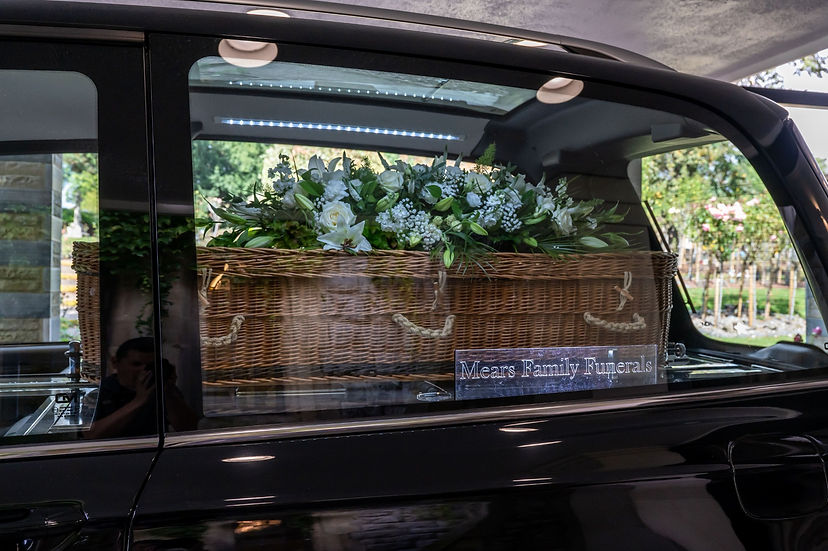 A wicker coffin with a white floral spray visible in the back of a hearse.
