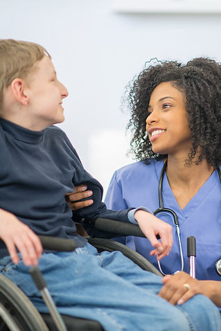 A nurse in blue scrubs smiles warmly at a person in a wheelchair