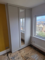 A white wardrobe with mirrored doors stands beside a large window in a room with yellow walls and exposed wooden flooring