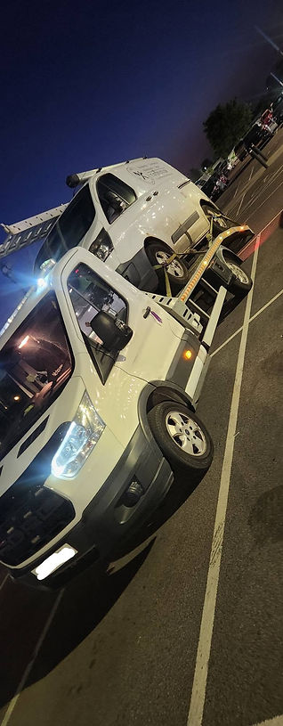 Side view of a white breakdown recovery truck carrying a silver car at night
