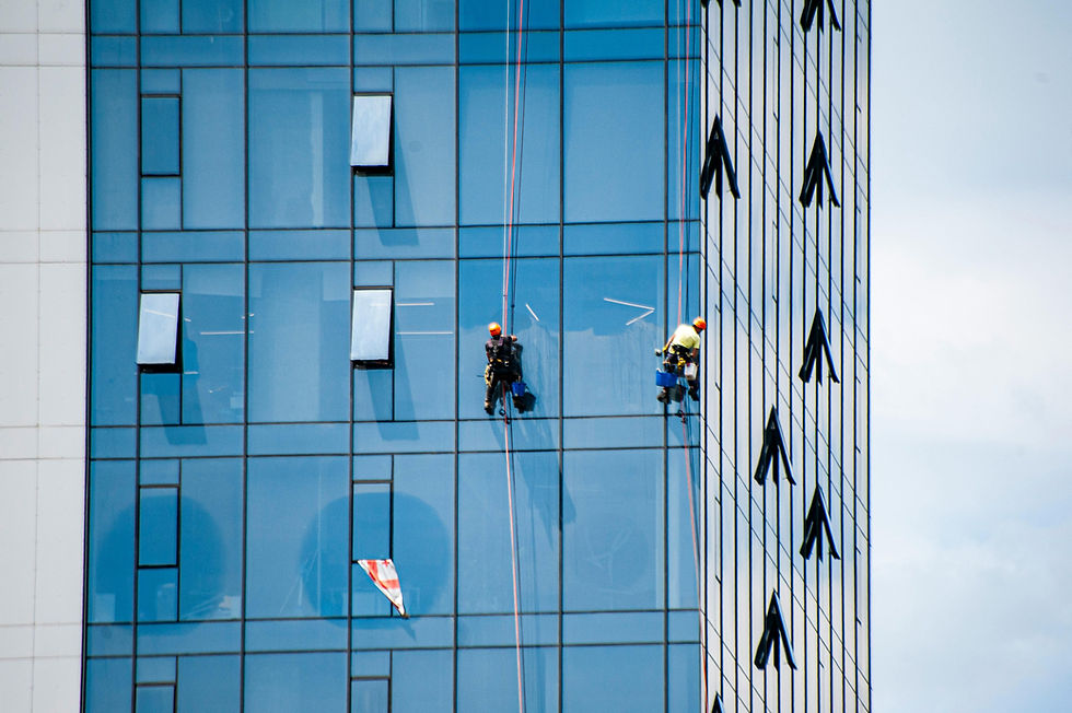 Window washers clean a blue glass skyscraper. Open windows and ropes visible. Bright sky background adds contrast.