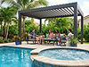 Family relaxing with a dog under a dark metal pergola on patio furniture near a pool.