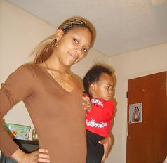 Adult wearing a brown top and gold jewelry holds a young child dressed in a red outfit with white patterns. They are in a residential room with light-colored walls and a wooden door in the background, which has a photograph taped to it. The image captures a moment of close bonding, possibly between family members.