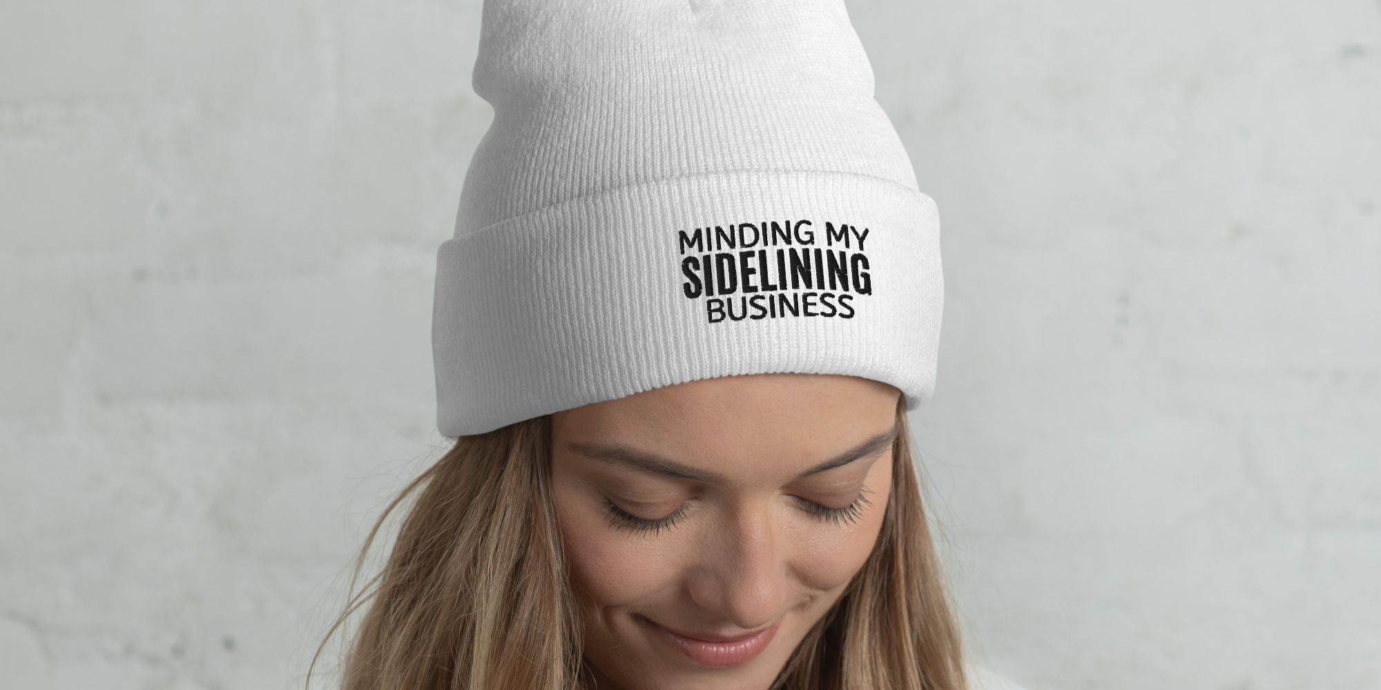 A person with long curly hair wears a white beanie featuring the printed phrase "MINDING MY SIDELINE BUSINESS." Paired with a