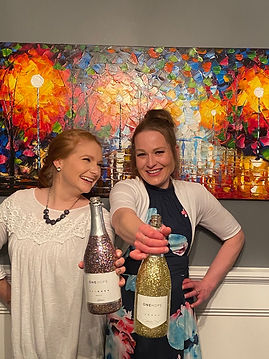 Two smiling women hold sparkling wine bottles labeled "ONEHOPE" in front of a colorful abstract painting. Festive and joyful atmosphere.