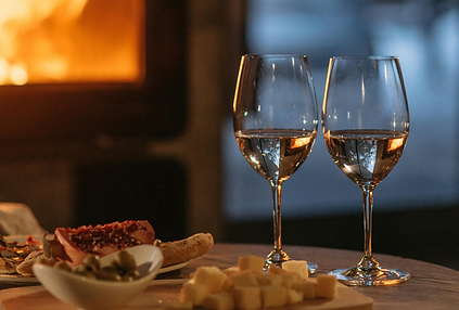 Two wine glasses on a table with cheese, bread, and a cut pomegranate. A cozy fireplace glows warmly in the blurred background.