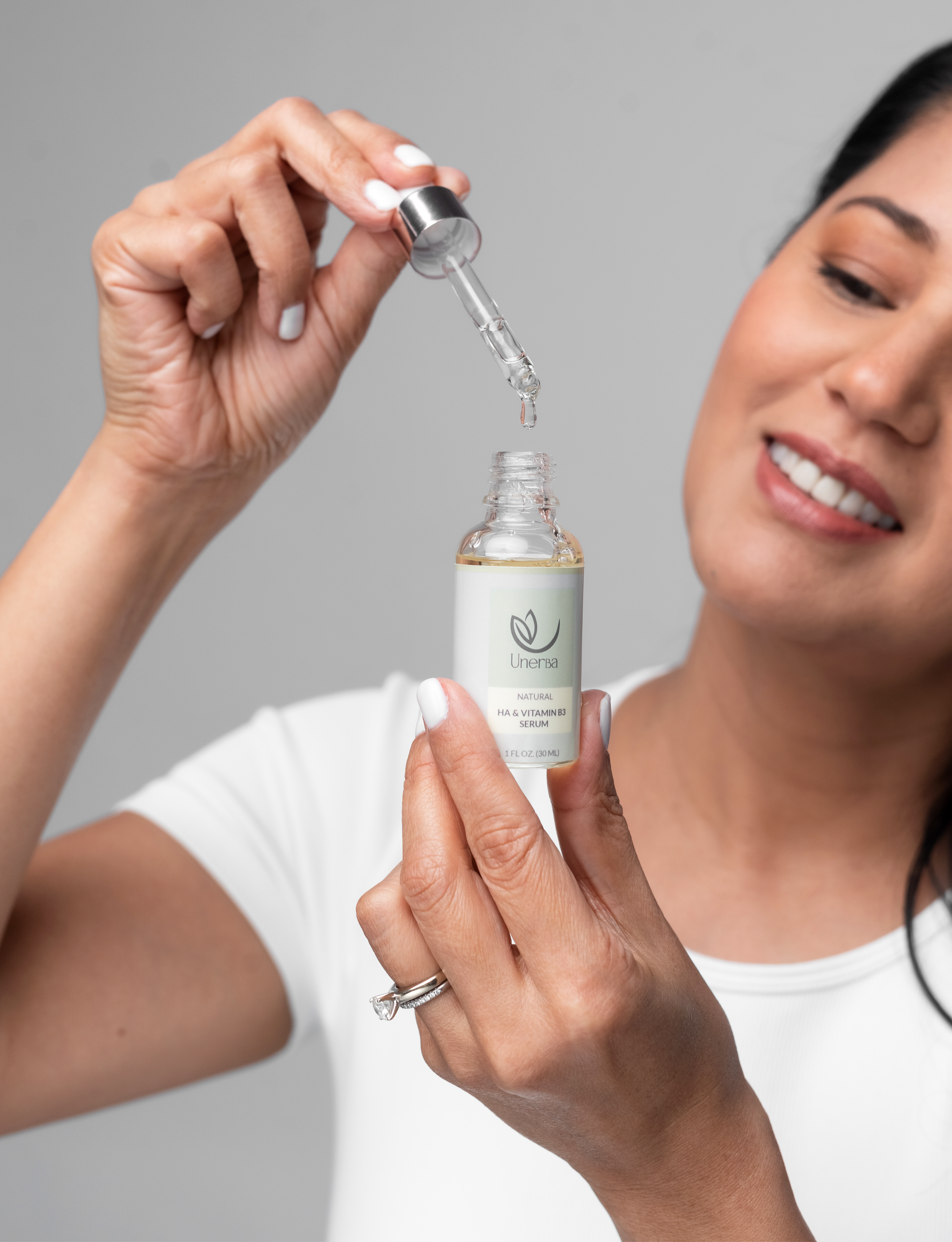 Model smiling holding the Hyaluronic Acid & Vitamin B3 bottle with her left hand while she is looking at the dropper dripping some of the product.