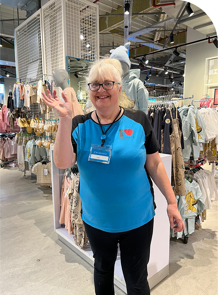 Woman in a blue "I ❤️ PRIMARK" shirt smiles and waves in a clothing store, surrounded by racks of children's clothes and mannequins.