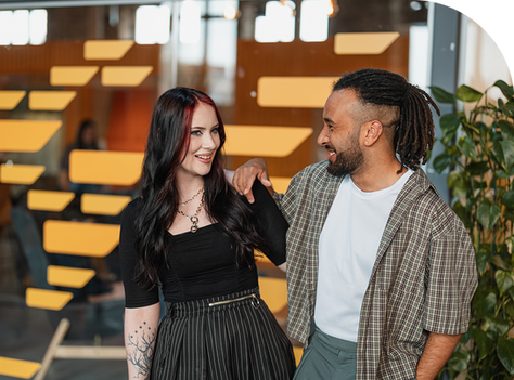 Two people smiling at each other in a modern office with orange decor. One has a tattoo, and the other wears a plaid shirt. Warm atmosphere.