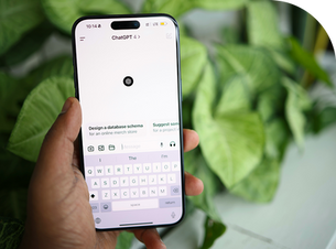 Hand holding a smartphone displaying the ChatGPT app, keyboard visible. Background shows green leaves, creating a calm setting.