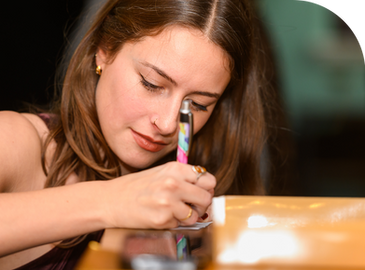 A woman with brown hair writes on paper with a colorful pen, focused expression. Blurred background, warm lighting.
