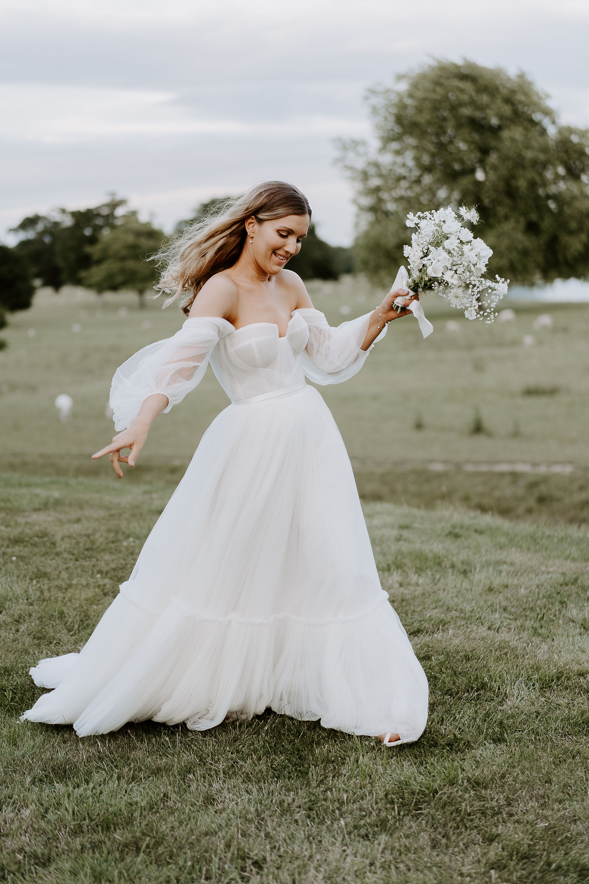 A bride in a white gown walks through a field holding a bouquet with white flowers. Modern bridal makeup by Emily Chantal.