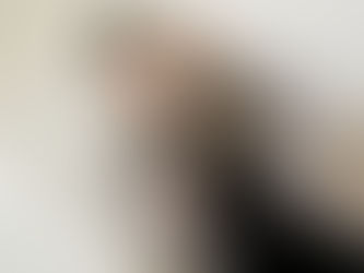 Emily Chantal, Suffolk and Essex makeup artist, sits in a bright room, holding makeup brushes. She touches her hair, looking content. Vanity with lights in the background.