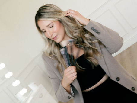 Emily Chantal, Suffolk and Essex makeup artist, sits in a bright room, holding makeup brushes. She touches her hair, looking content. Vanity with lights in the background.