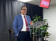 Pastor speaking behind a clear podium at Compassion Church Albuquerque with black drapery, greenery, and church branding in t