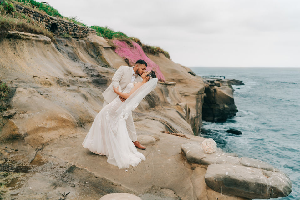 groom dips bride on La Jolla cliff with ocean and purple flowers behind them.