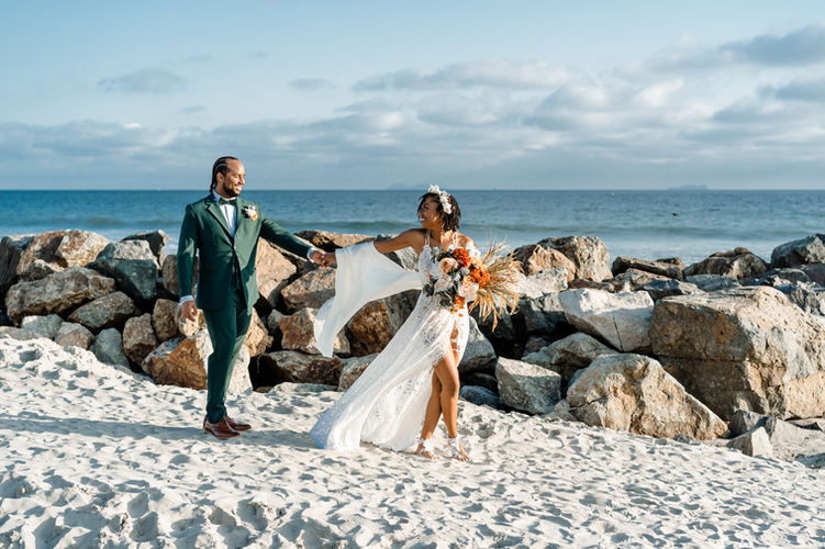 bride leading groom in the sand with the ocean and rocks in the background.