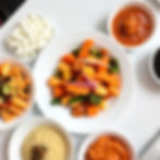 A white plate of sautéed or steamed vegetables (carrots, potatoes, and green peppers) surrounded by small bowls of cheese chunks and various side dishes and sauces.