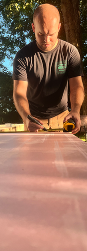 Trent a bald man in a black shirt with a tape measure and marker measuring pink foam board