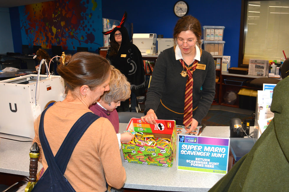 Woman in costume assists a mother and child at a library counter. "Super Mario Scavenger Hunt" sign and bright comic-patterned box visible.