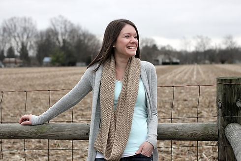 Smiling woman standing in front of a fence with a field and trees in the background.