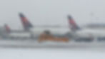 Snow-covered airport with several airplanes parked. A snowplow clears the runway amidst falling snow, creating a cold and busy atmosphere.