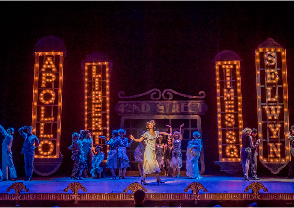 A theatrical stage scene with a central performer and a backdrop of illuminated vintage theater signs.