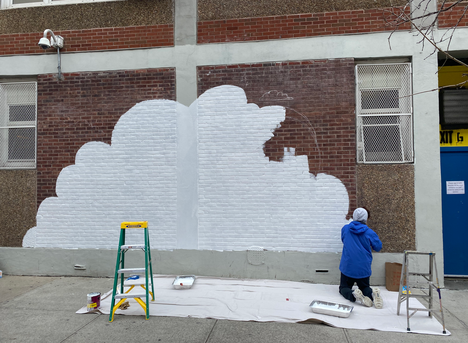 Photo from behind of a person in a blue jacket kneeling at the lower right corner of a wall, priming for a mural. The wall depicts a large white cloud, almost completely painted.