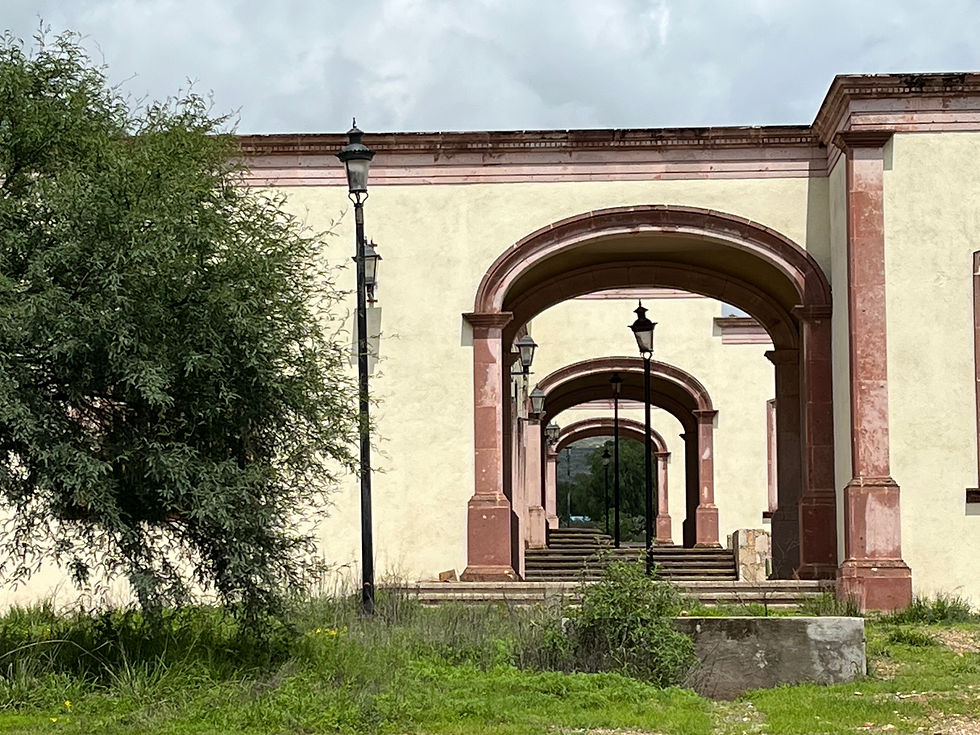 Arched stone corridor with lamp posts and pink trim, leading to a garden. Greenery and a tree on the left under a cloudy sky.
